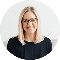 A person with long, straight blonde hair and glasses smiles while looking at the camera. They are wearing a black top and are seated against a plain white background.