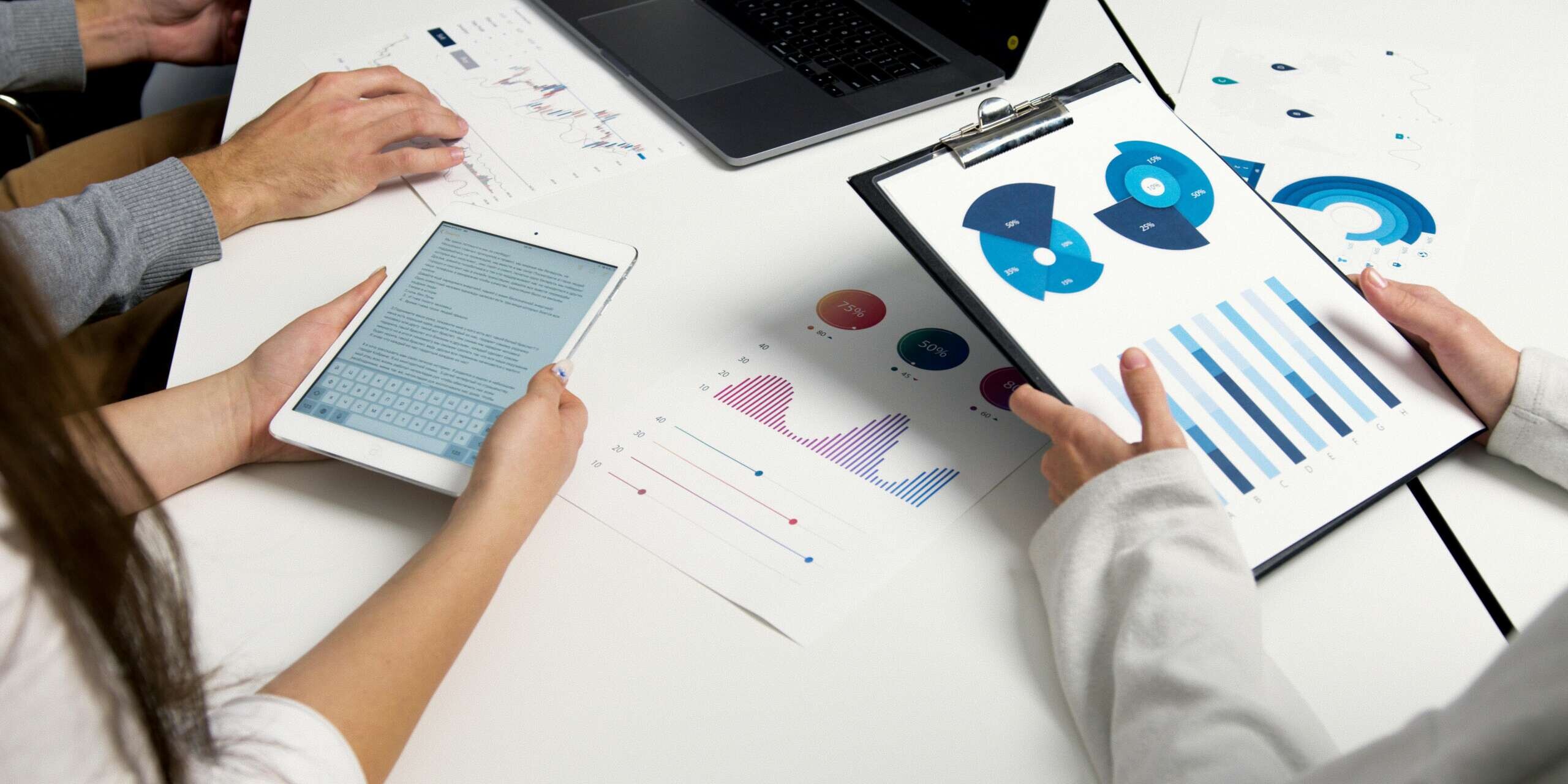 People collaborating at a table with charts, graphs, a tablet, and a laptop. One person is holding a clipboard with a pie and bar chart, while another types on a tablet. Various printed graphs are spread on the table.