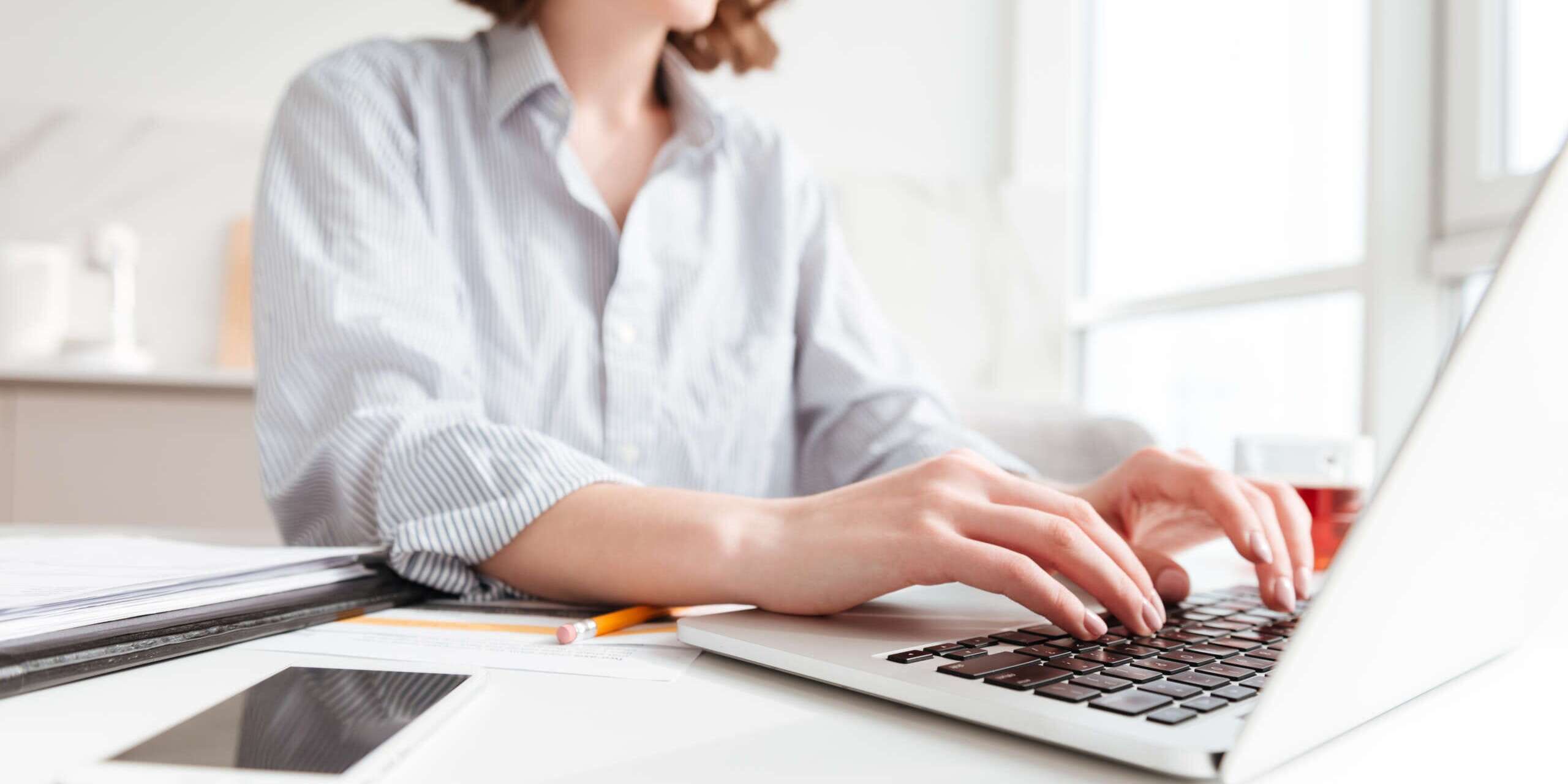 A woman in a striped shirt is smiling while typing on a laptop at a white desk. The desk also has a smartphone, a pen, and a notebook. Sunlight streams through a window in the background.