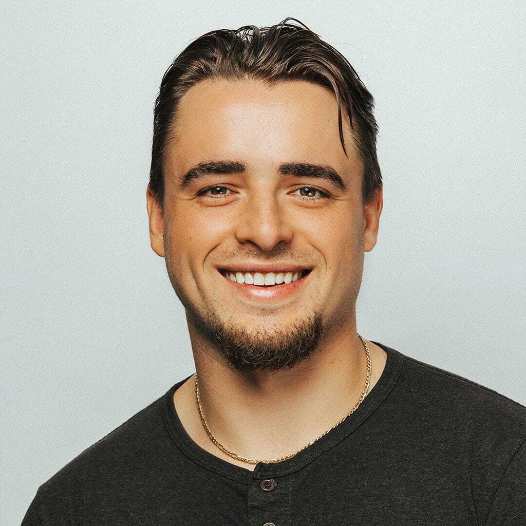 Smiling person with short brown hair and facial hair, wearing a dark shirt and a gold chain necklace against a plain light background.