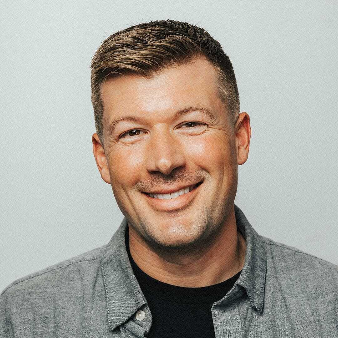 A handsome smiling man with short brown hair wears a gray button-up shirt over a black T-shirt. The background is plain and light-colored.