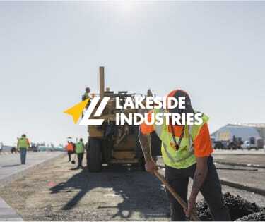 A construction worker in a high-visibility vest is shoveling material on a sunny day with machinery and other workers in the background. The image has a logo and text that reads Lakeside Industries.