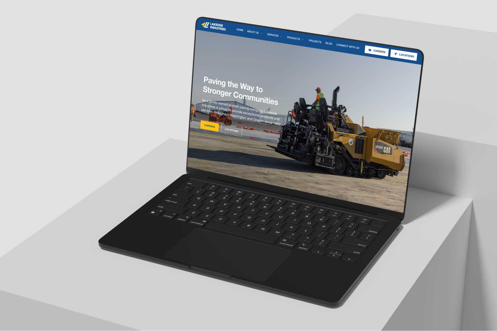 A laptop displaying a construction companys website homepage. The screen shows workers operating heavy machinery on a road with the text Paving the Way to Stronger Communities. The site has a dark blue header with a navigation menu.