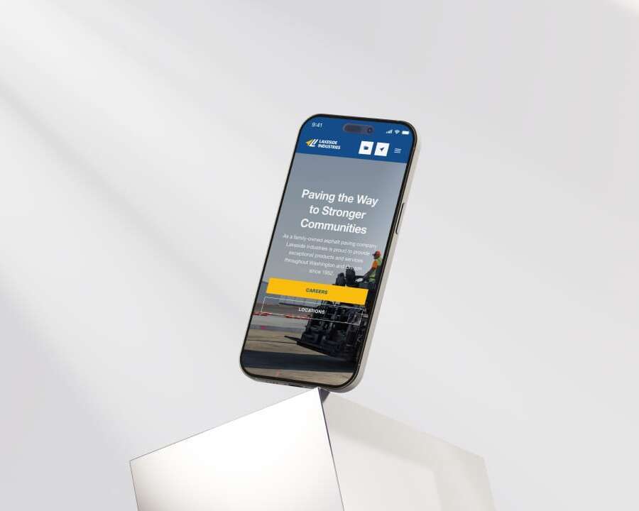 Smartphone displaying a website with the text Paving the Way to Stronger Communities, featuring construction imagery. The phone is propped on a reflective surface against a minimalist background.