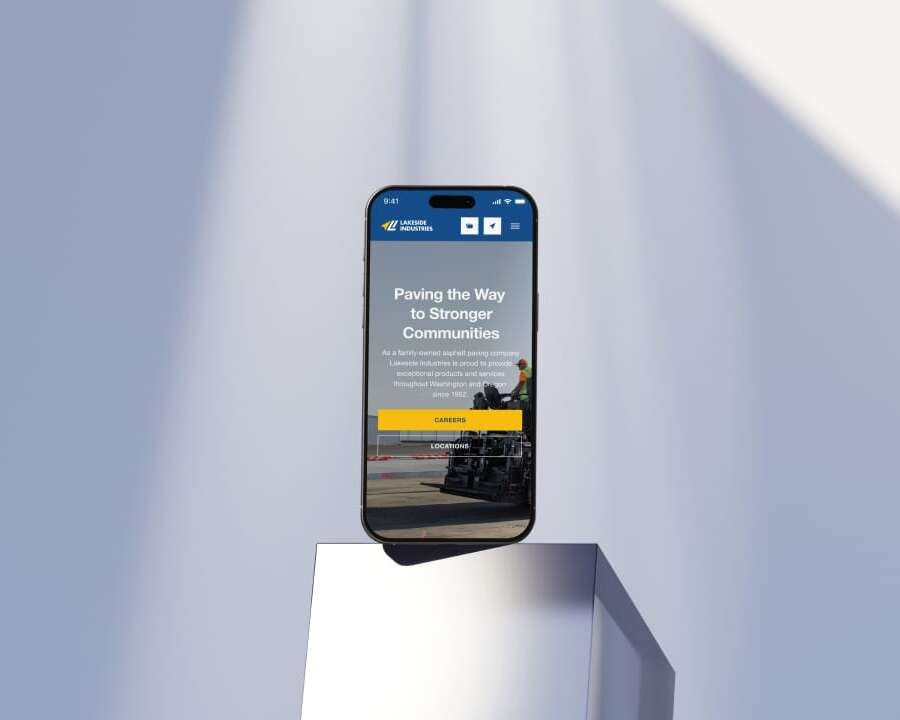 A smartphone displaying a website stands on a metallic pedestal, bathed in soft light. The website has a blue header with icons, and text that says Paving the Way to Stronger Communities alongside an image of construction work.