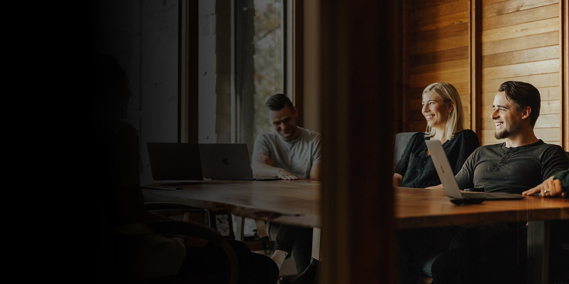 Three people are sitting around a wooden table in a conference room with laptops open. Two are smiling, and a window lets in natural light, illuminating the room with wood-paneled walls.