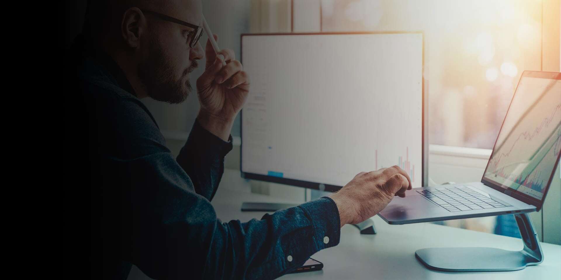 A person with glasses works at a desk, holding a pen while looking at a laptop. A monitor displaying graphs is next to them, and sunlight streams through the window. The room has a modern, professional atmosphere.
