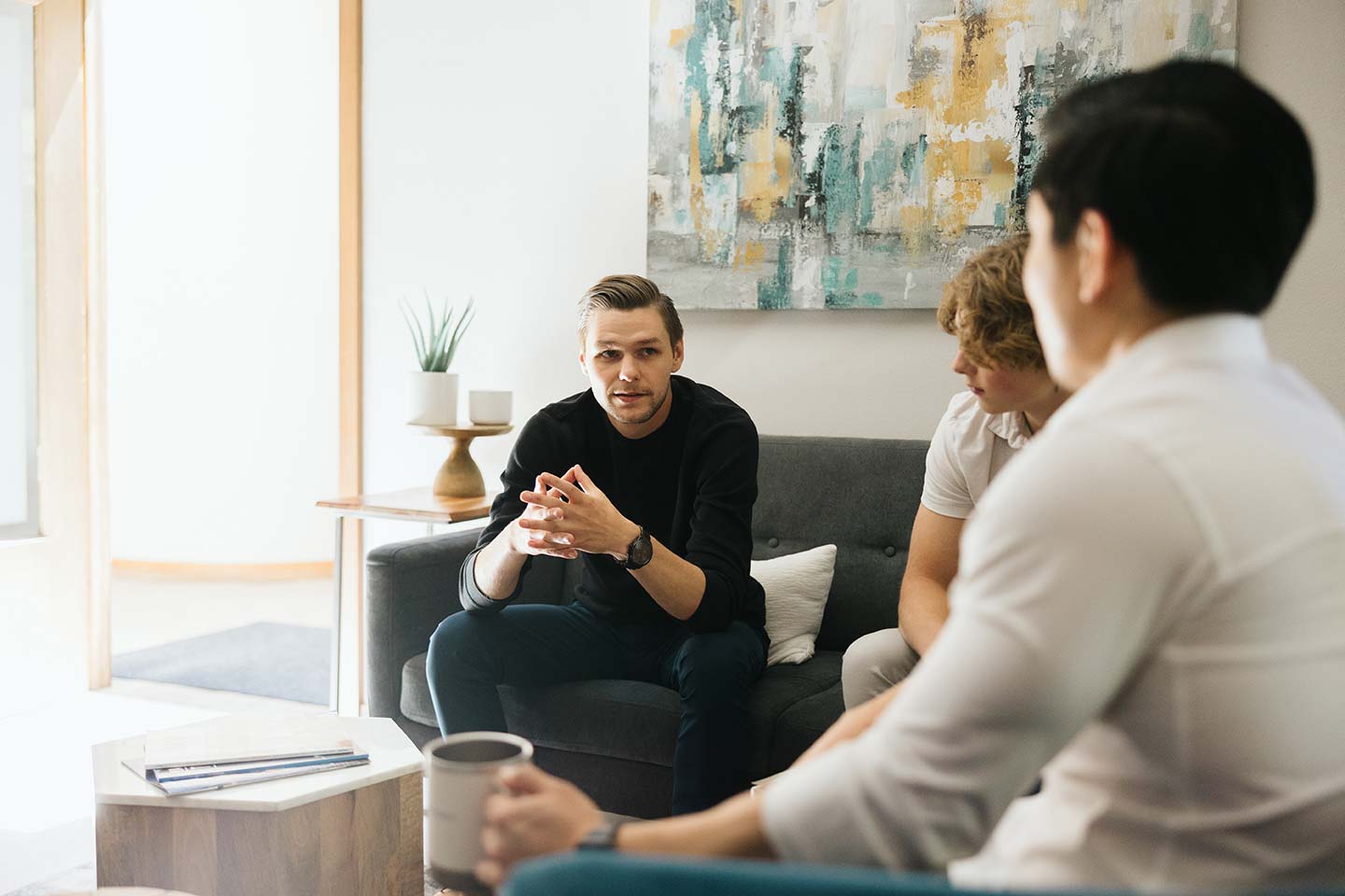 Three people sitting on a couch in a living room, engaged in conversation. A young man with a watch is speaking, while another person attentively listens. A third person holds a coffee mug. A modern painting and small plant decorate the room.
