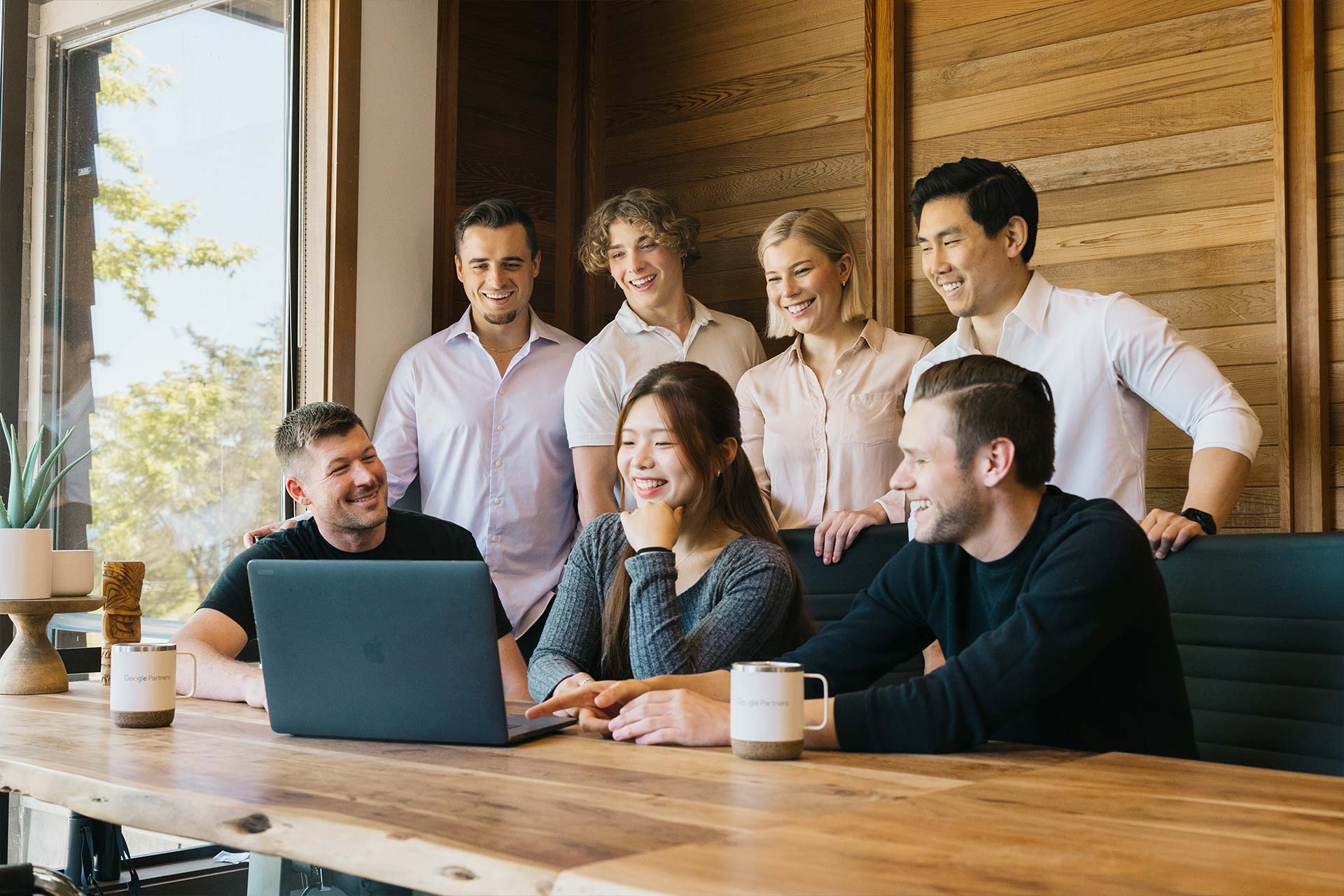 A group of seven people gather around a laptop on a wooden table, smiling and engaged. Coffee mugs are on the table, and a large window with a view of trees is in the background. The setting appears to be an office or meeting room.