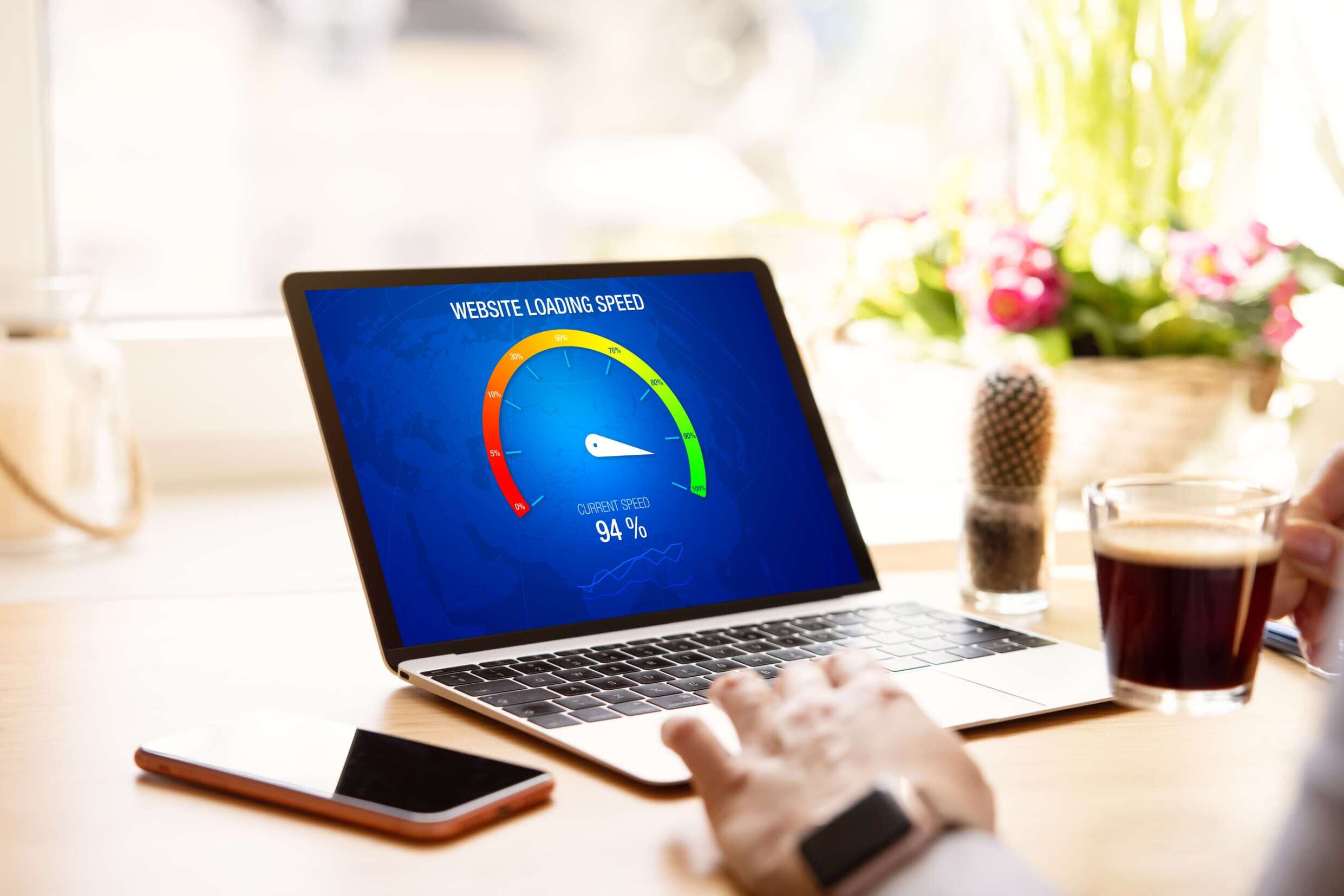 A person is using a laptop showing a website loading speed graph at 94% on the screen. The scene includes a cup of coffee, a smartphone, and a small plant in the background on the wooden table. Bright light filters through a nearby window.