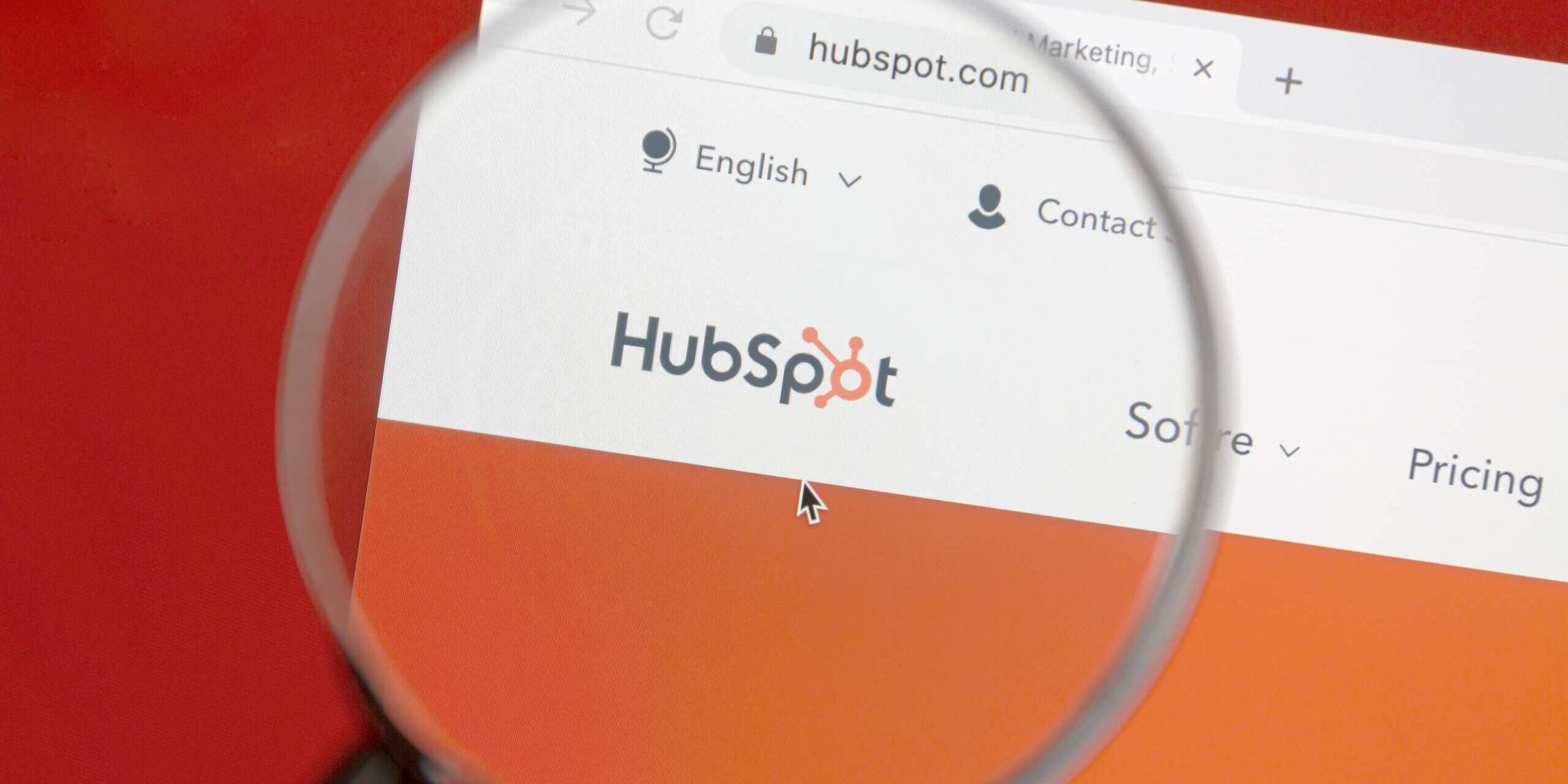 A computer screen displaying the HubSpot website under a magnifying glass. The browser tab shows navigation options like Software and Pricing on a predominantly orange background, focusing on the HubSpot logo.