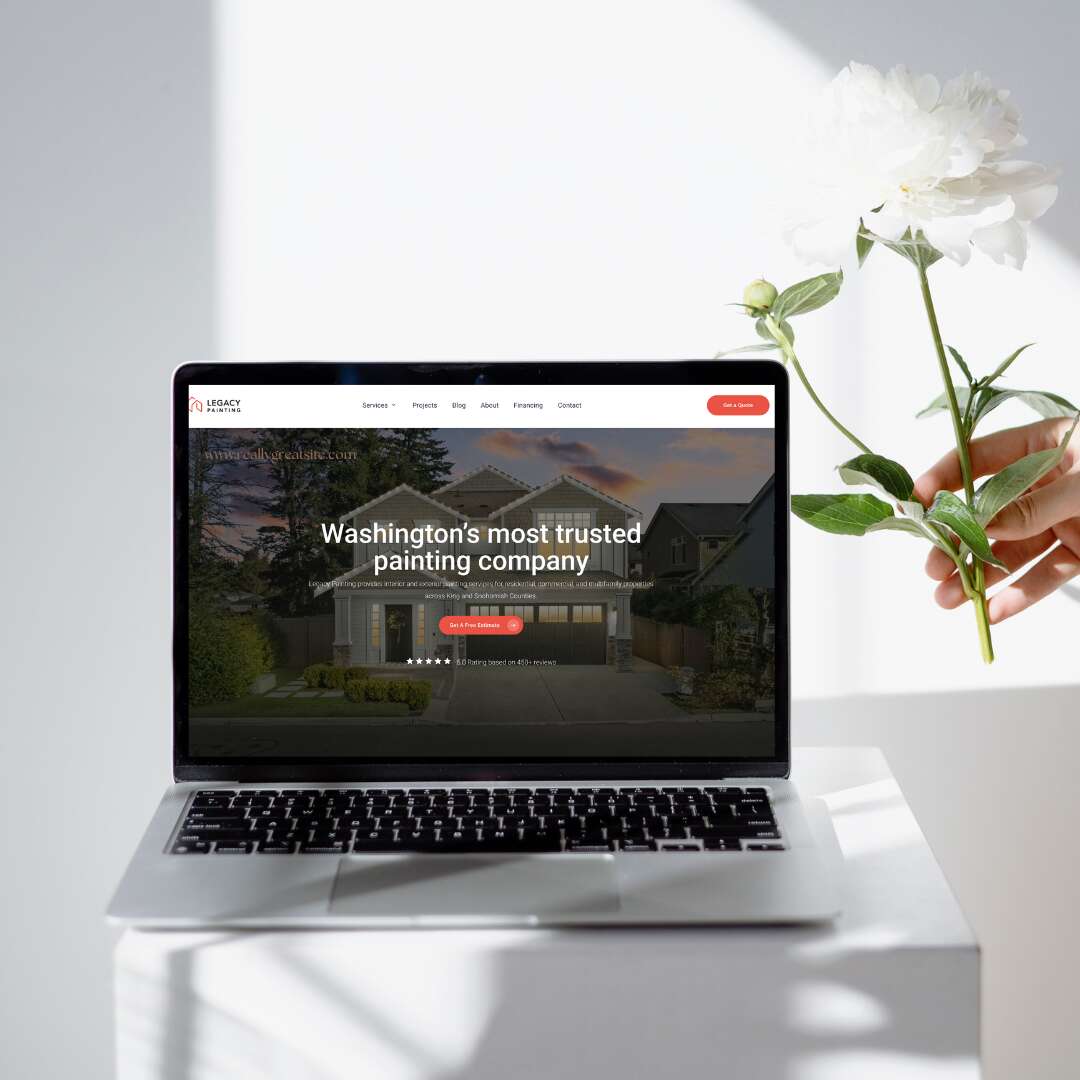 A laptop displaying a painting companys website sits on a white table. A hand holds a white flower beside the laptop, and soft sunlight casts shadows on the wall and table.