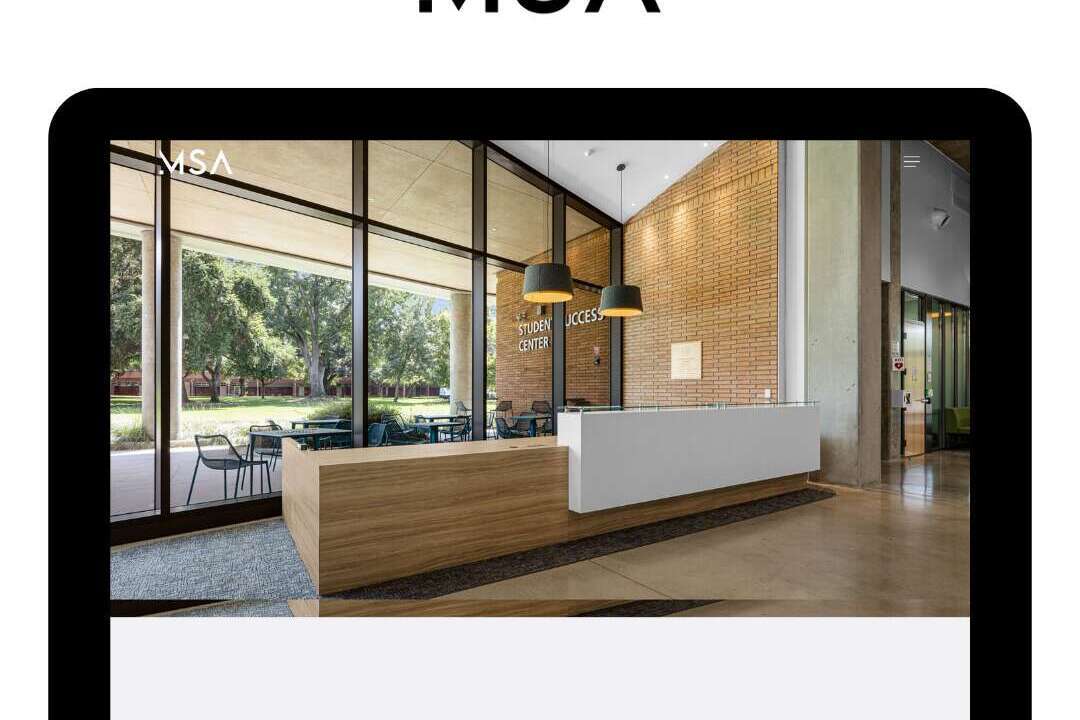 A modern office reception area with a sleek wooden desk, white panel, and large windows letting in natural light. The image is displayed on a device screen with the MSA logo above it.