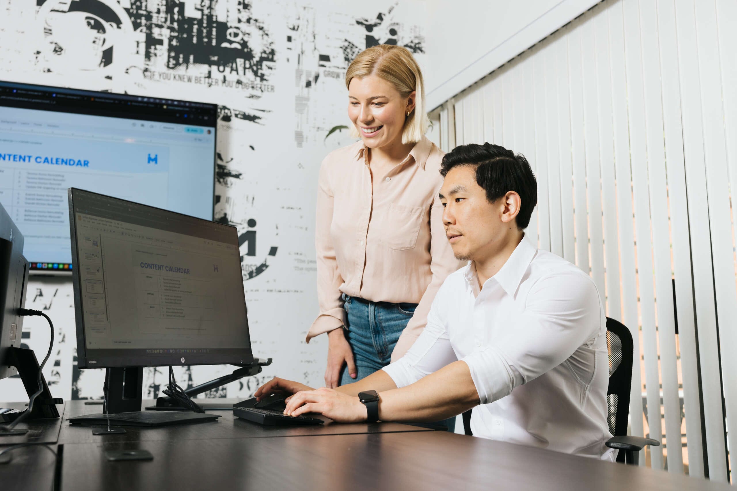 A man sits at an office desk, focused on a computer screen displaying a content calendar. A woman stands beside him, smiling and looking at the screen. The room has a modern design with a graphic wall and vertical blinds.