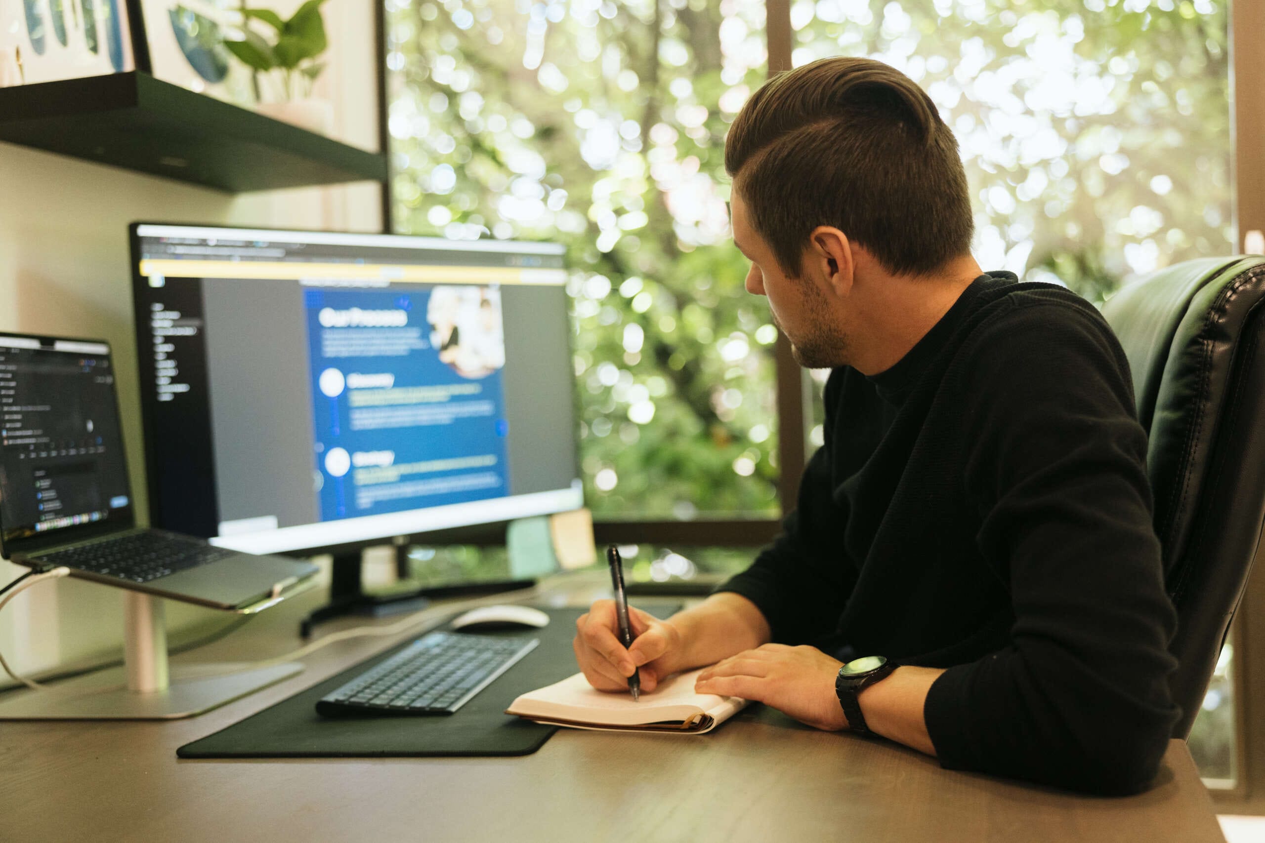 A person sits at a desk with a notebook and pen, writing while viewing a large computer monitor. The room has a large window with leafy greenery outside. Shelves with books are visible. The person wears a black sweater and a watch.