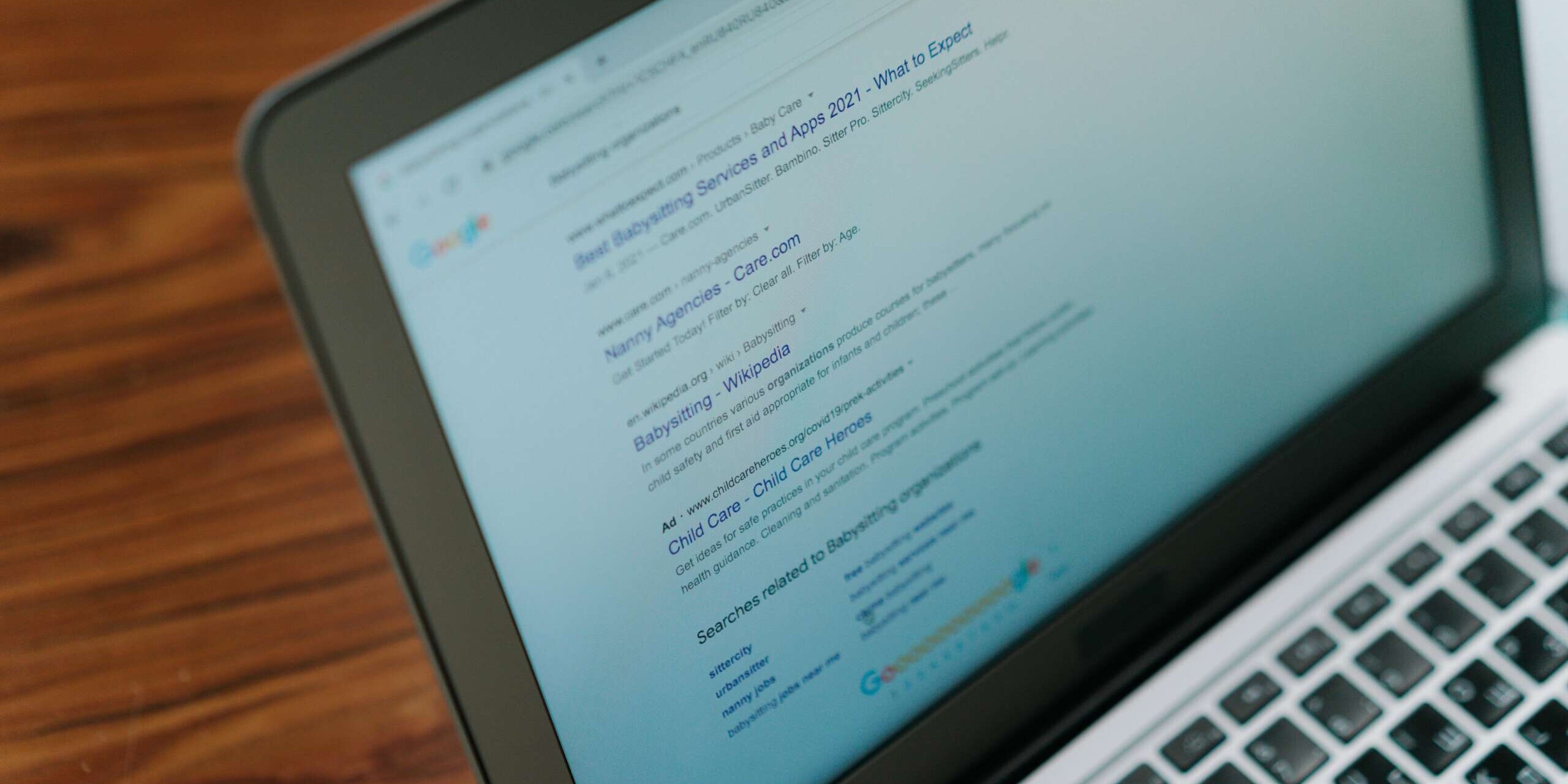A laptop on a wooden table displays a Google search results page about babysitting services and agencies. The screen shows multiple links with titles and brief descriptions related to the topic.