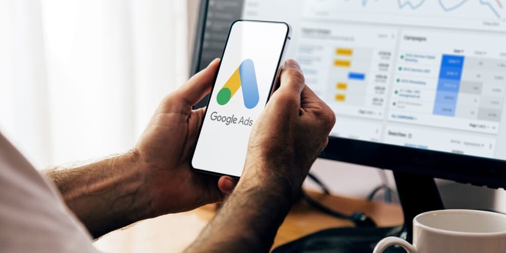 Person holding a smartphone displaying the Google Ads logo. In the background, a computer monitor shows analytics graphs and data related to advertising campaigns. A white coffee mug sits on the desk.