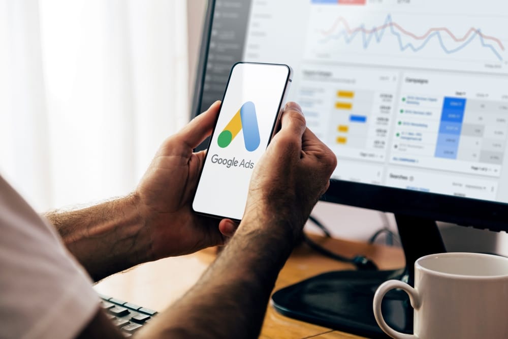 Person holding a smartphone displaying the Google Ads logo. In the background, a computer monitor shows analytics graphs and data related to advertising campaigns. A white coffee mug sits on the desk.