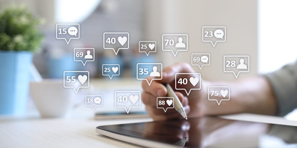 A person uses a tablet with a stylus, surrounded by floating social media icons indicating likes, comments, and followers in varying numbers. A potted plant is blurred in the background.