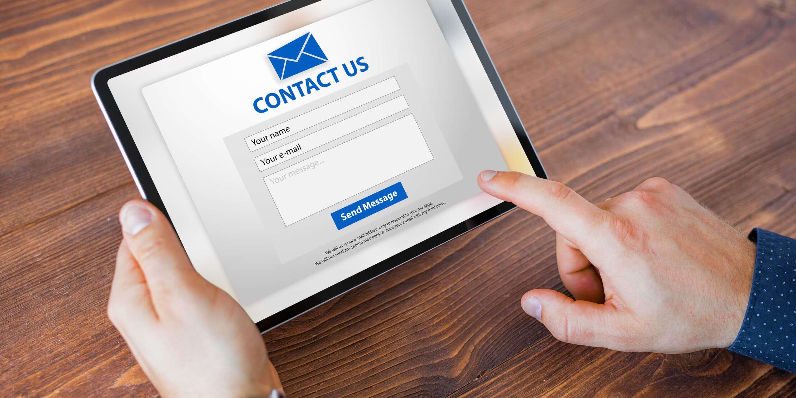 Person holding a tablet displaying a Contact Us form on a wooden surface. The form includes fields for name and email, with a blue send button at the bottom.