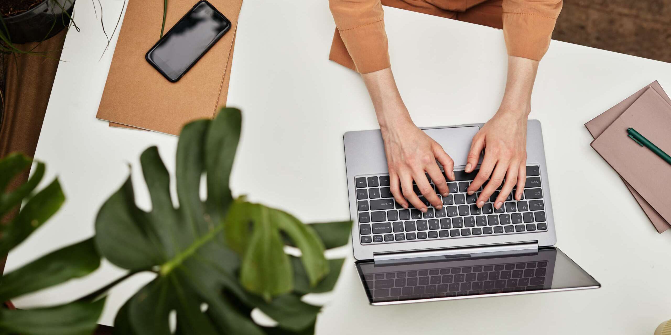 A person types on a laptop at a white desk with a smartphone, brown folder, and a pen. Green plants partially frame the scene, creating a natural and cozy workspace atmosphere.