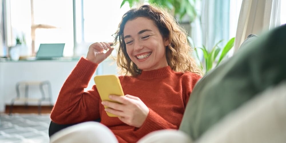 Smiling person with curly hair and a red sweater looking at a yellow phone while sitting on a couch. A blurred background shows a bright room with plants and large windows.