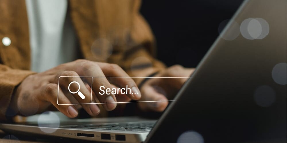 Person typing on a laptop with a transparent search bar overlay, featuring a magnifying glass icon next to the word Search.... The background is blurred, focusing on the hands and keyboard.