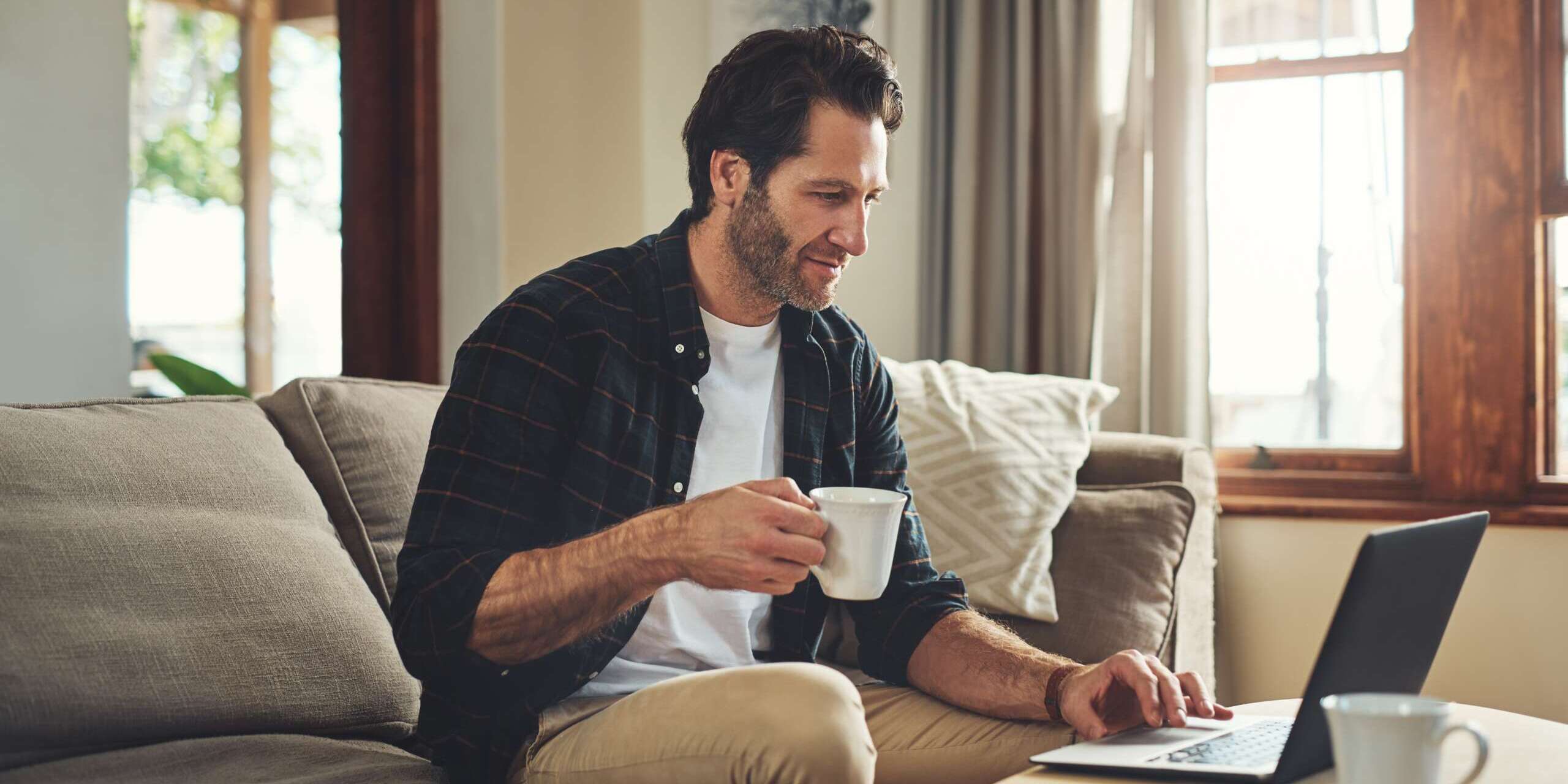 A man sits on a sofa, holding a coffee mug and using a laptop on a small table. Theres a cup and smartphone beside him. The room is bright with sunlight coming through a window, and the decor is cozy and modern.