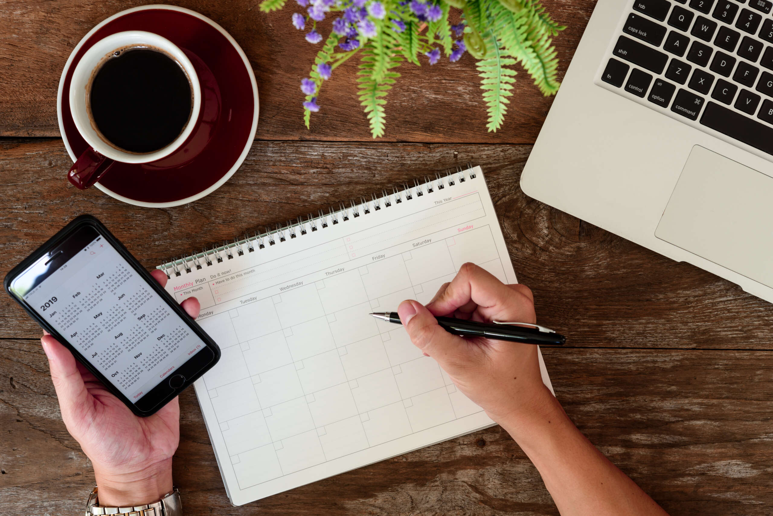 A person writes on a calendar with a pen, holding a smartphone displaying a calendar in the other hand. A laptop and a cup of coffee sit on a wooden table, with a small plant in the corner.