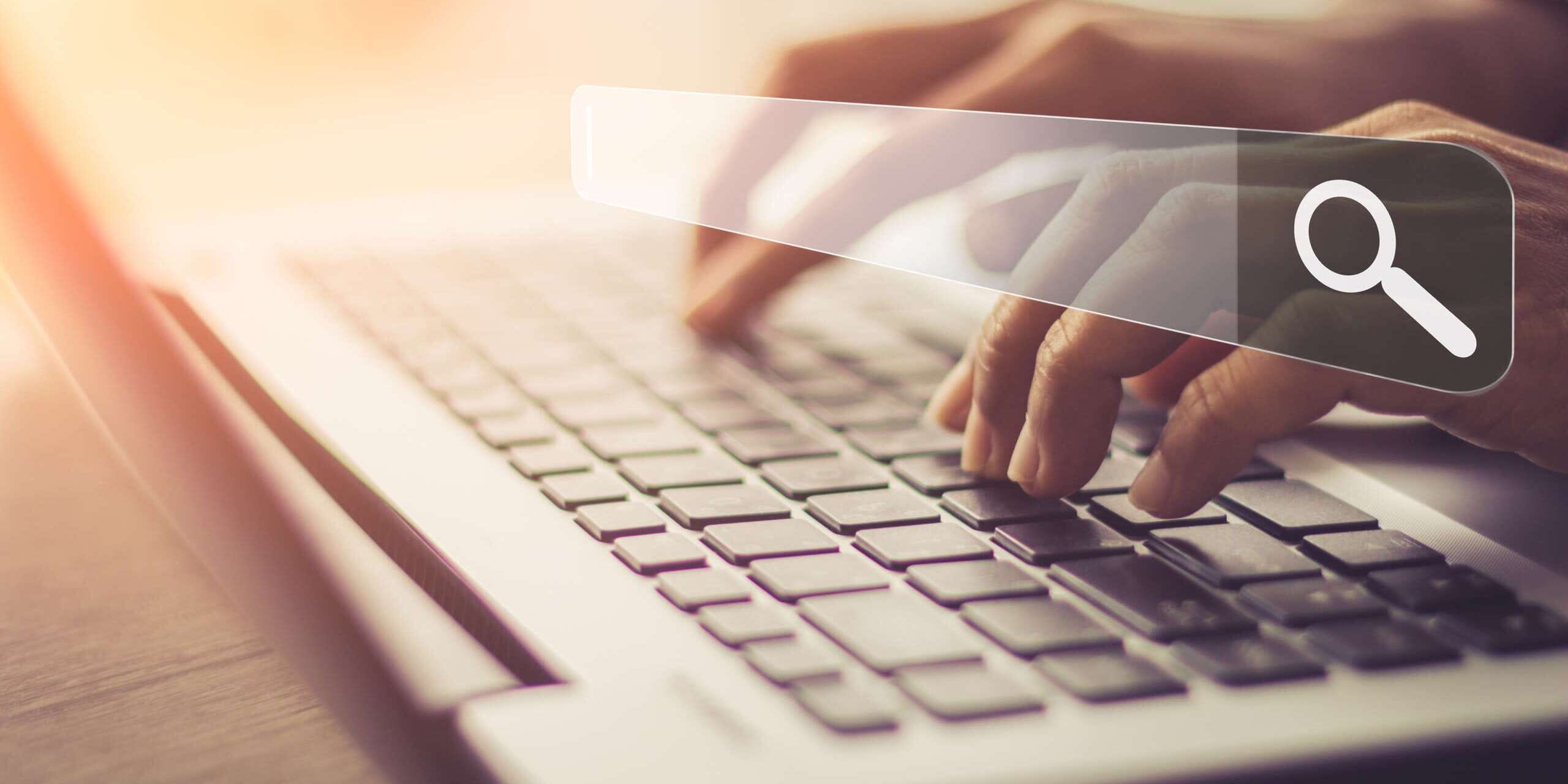 Close-up of hands typing on a laptop keyboard, with a transparent search bar and a magnifying glass icon overlaid above the keys, symbolizing searching or browsing online. The background is softly lit with a warm hue.