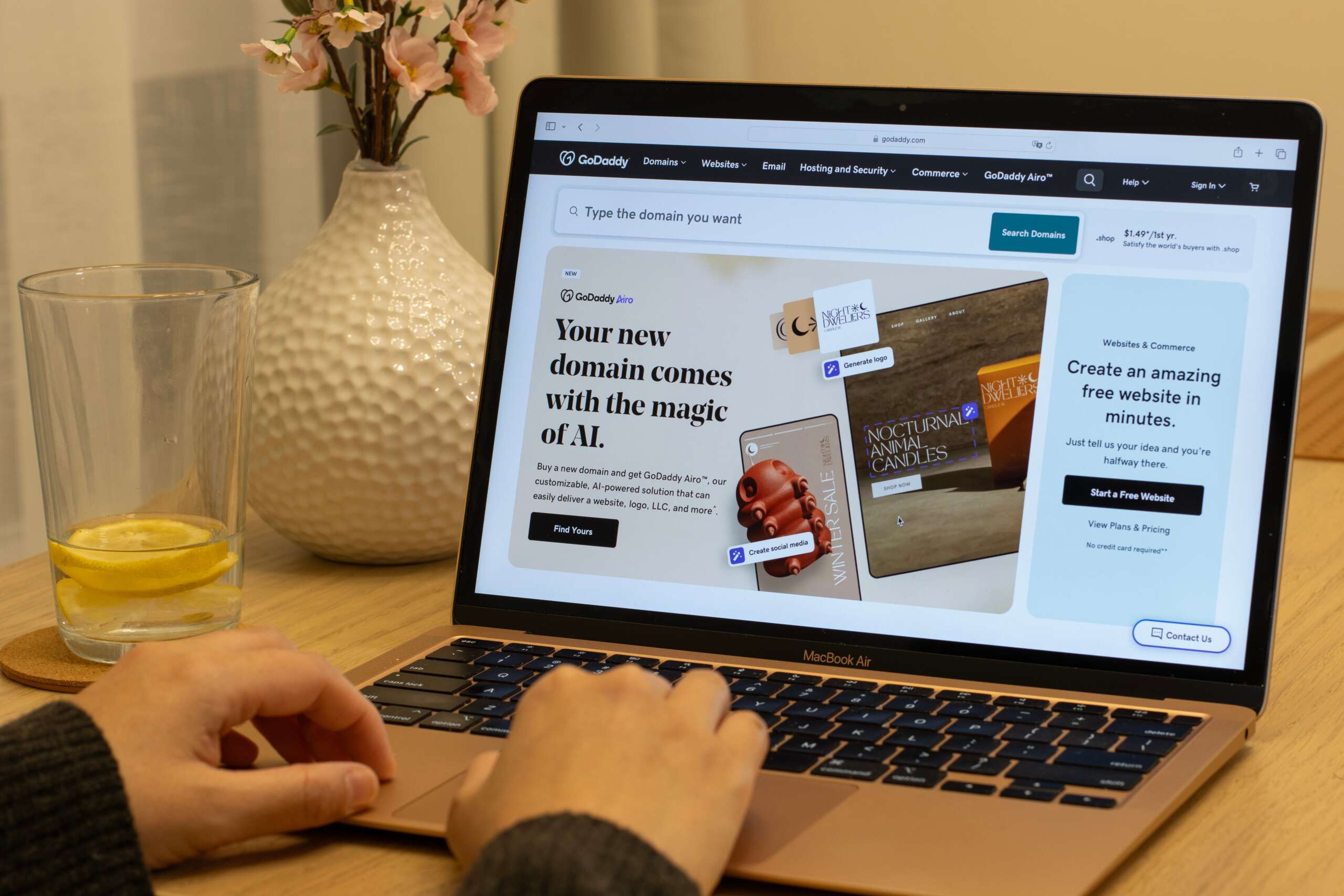 A person is using a laptop displaying a GoDaddy webpage about domain registration. The screen features promotional text and images. A glass with a lemon slice and pink flowers in a vase are on the wooden table.