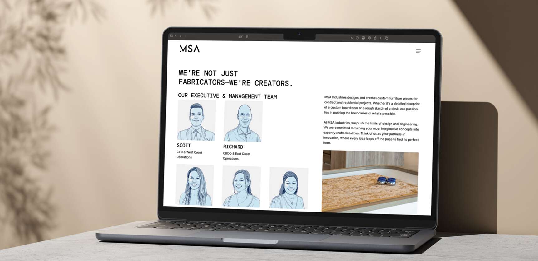 A laptop on a desk displays a website for MSA, showing sketched portraits of executives named Scott and Richard, a team description, and an image of a wooden project. Sunlight casts shadows on the desk.