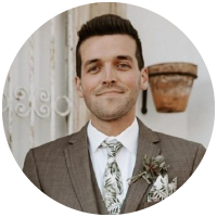 A man in a brown suit with a white shirt and patterned tie, smiling, stands in front of a rustic wall and a hanging flowerpot. He has short dark hair and is wearing a boutonniere.