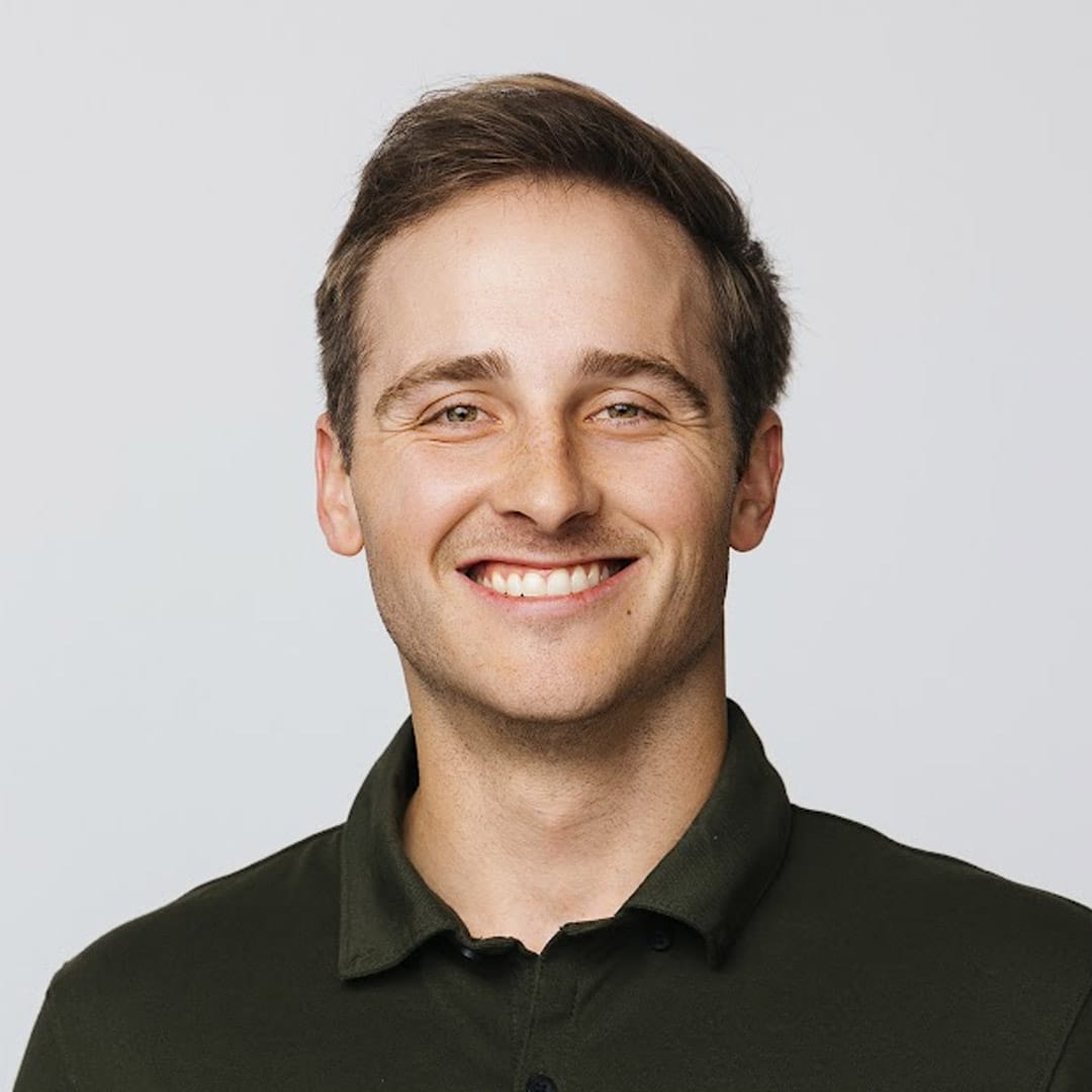 A young man with short brown hair smiles at the camera. He is wearing a dark green collared shirt and standing against a plain, light-colored background.