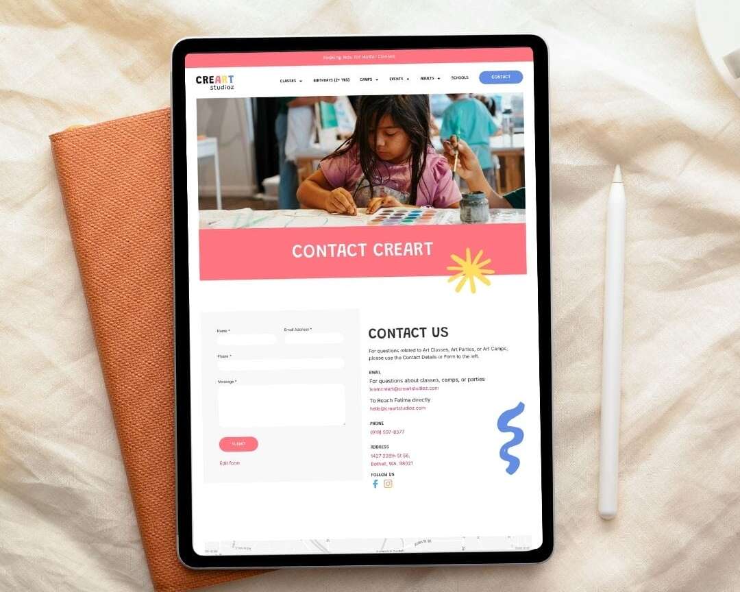 A tablet on a white fabric surface displays a website contact page with a photo of a child painting, a contact form, and a section labeled CONTACT CREART. A closed notebook and a stylus are beside the tablet.