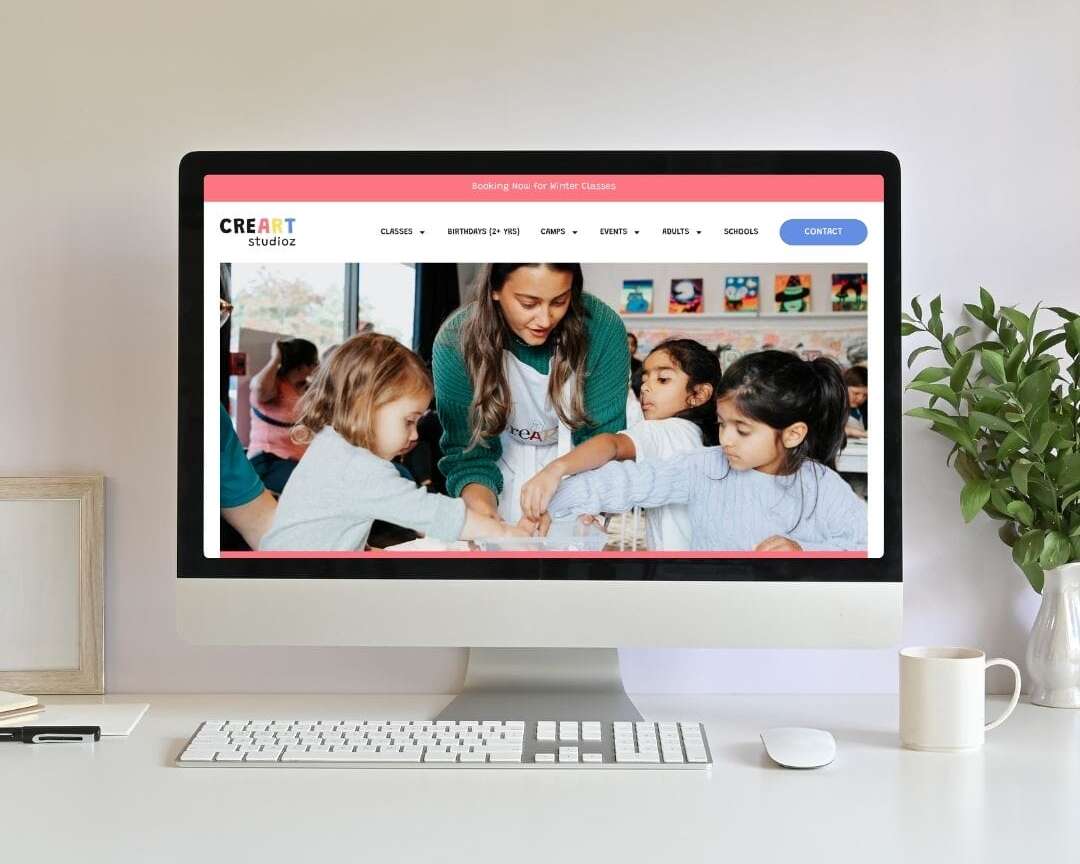 A computer on a white desk displays a website showing a group of young children doing an art activity with an adult. The desk has a plant, framed picture, keyboard, and mouse.