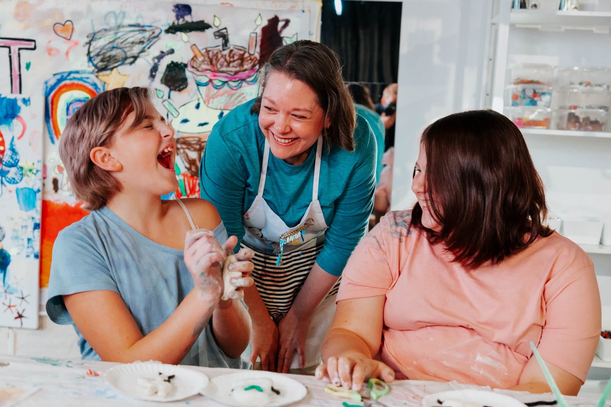 Three people sit at a table doing crafts, smiling and laughing. The background features a colorful, painted wall, suggesting a fun, creative environment. One person stands, engaging warmly with the other two.