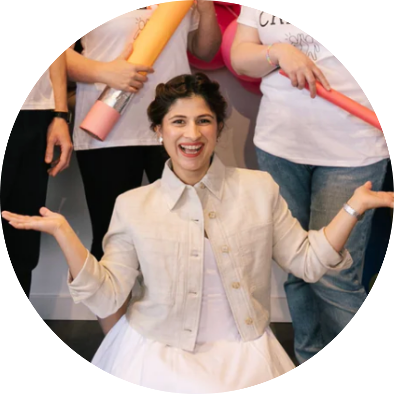 A woman in a light-colored jacket and white dress kneels, smiling with arms raised. Behind her, three people stand holding large colorful rolled papers, but their faces are cropped out of the frame.