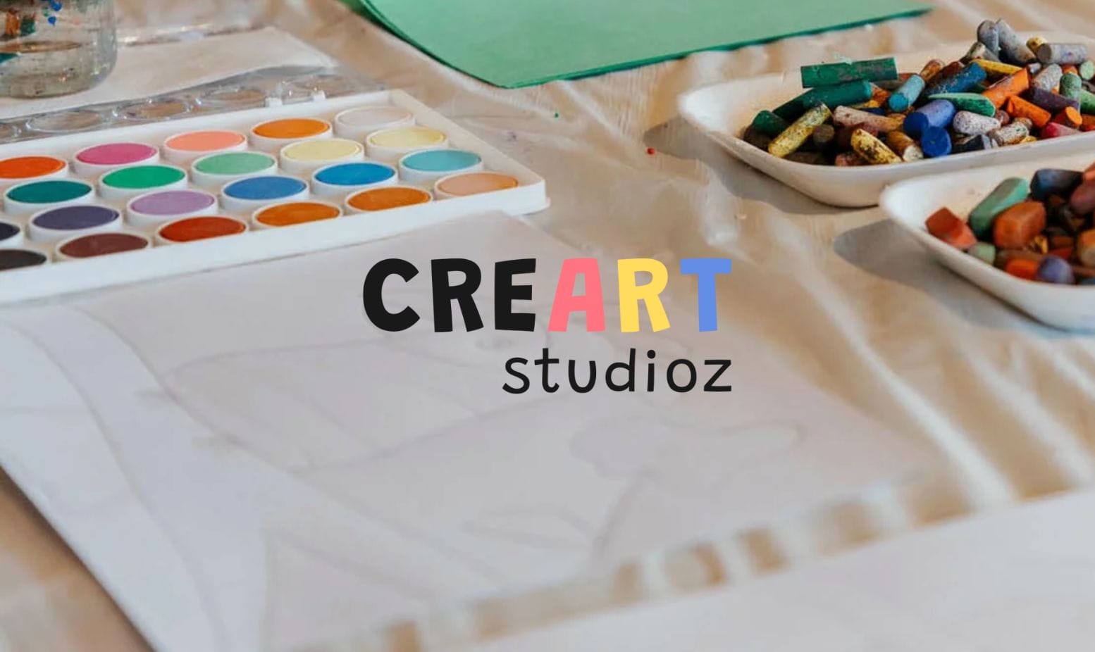 Art supplies including watercolor paints, colored chalks, and paper are arranged on a table with a faint pencil sketch. The words CREART studioz appear in the center, with ART in colorful letters.