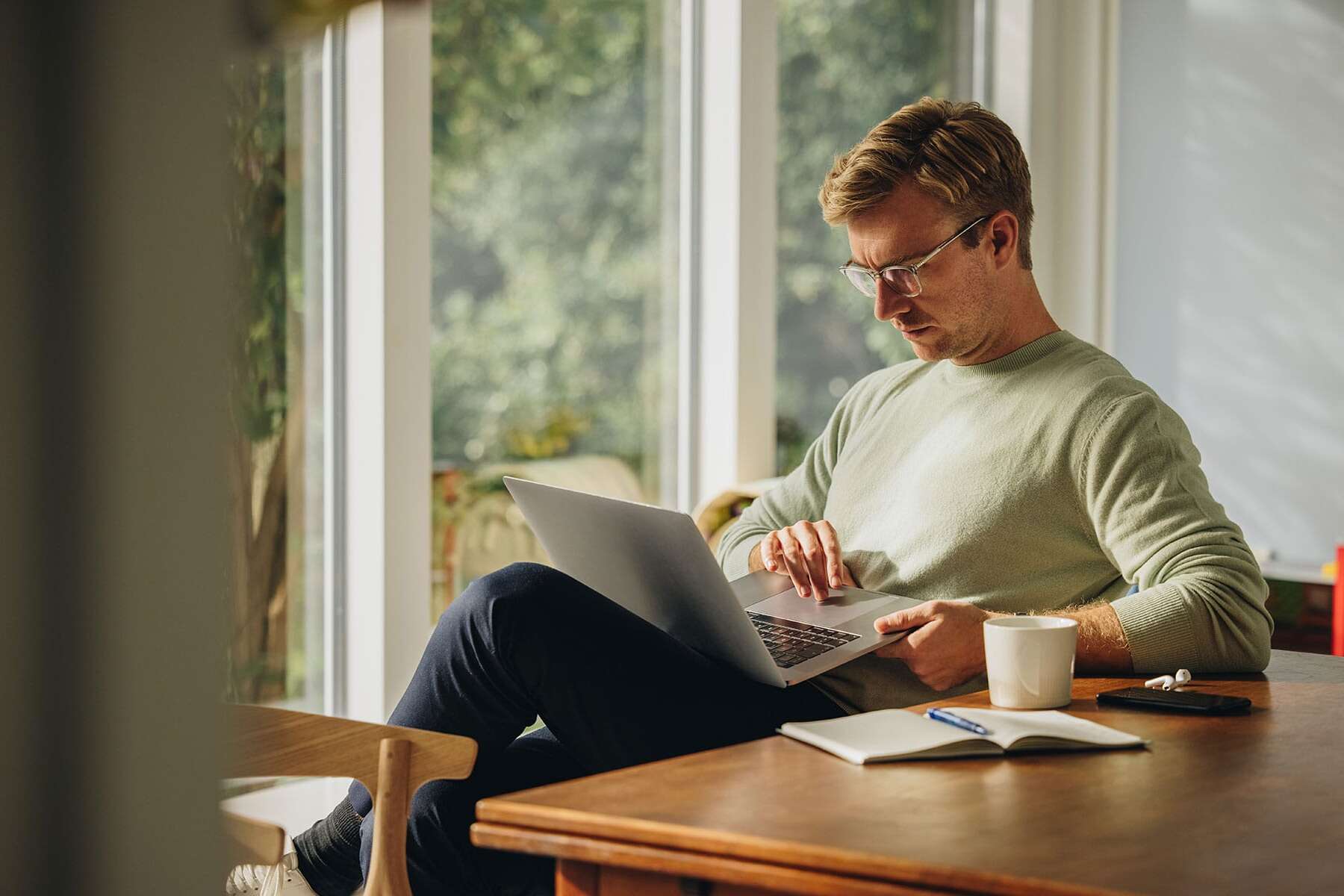 A person wearing glasses sits at a wooden table, using a laptop. There is a notebook, pen, and coffee mug on the table, with large windows and greenery visible in the background.